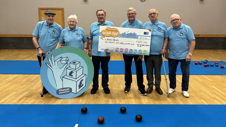 This photo shows six of the members of the group standing in a row holding a prop big cheque. In front of them is a blue mat with a set of bowls laid out on it. 