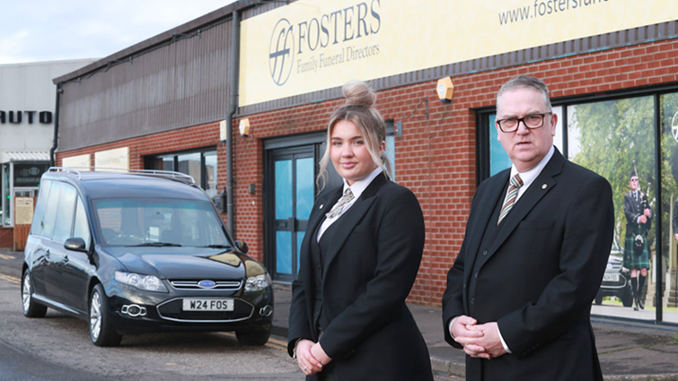 This photo shows Ellie and Funeral Manager John Brown, both in full uniform, standing in front of Fosters funeral care in Rutherglen and with a hearse parked in the background.
