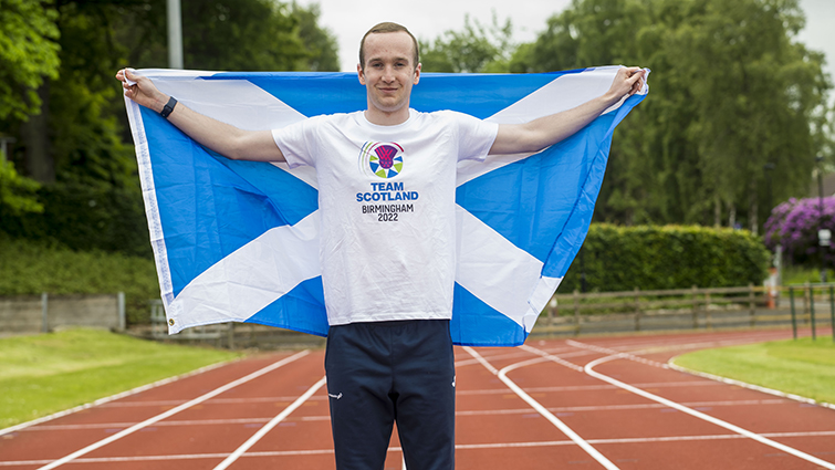 This photo shows para athlete Alexander wearing Team Scotland colours and holding up a Scotland flag. He is pictured on a running track.