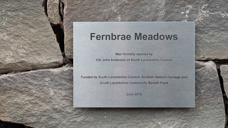 a square silver coloured metal sign on a  grey stone wall. The sign reads Fernbrae Meadows in bold text and has further details on when the park opened and the funding partners.