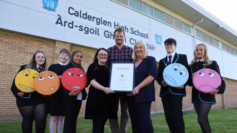 In this photo the Head Teacher and two depute head teachers are standing in the middle of a group of five school pupils in front of Calderglen High School. Each pupil is holding a colourful 'face' prop showing a different expression or mood.