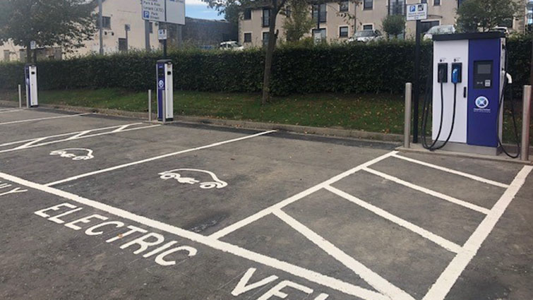 This is a picture of the Carluke Lifestyles Car Park to the right is an electric vehicle charging point in silver and blue. In the foreground are parking spaces marked for use by electric vehicles only.