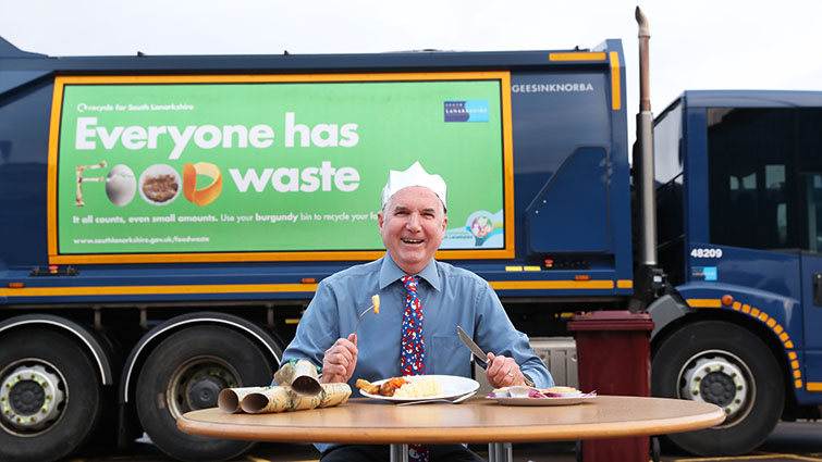 Councillor John Anderson, wearing party hat and Christmas tie sits at a small table tucking into a Christmas dinner, with one of the council's refuse vehicles bearing the food waste campaign advert, behind him.