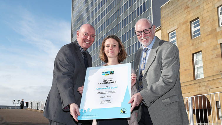 Chief Executive Cleland Sneddon, Food policy officer Helene Gourichon and Council Leader, John Ross are standing together on the steps of council headquarters with an enlarged version of the council's Fairtrade certification.