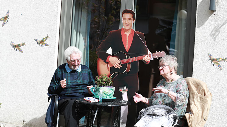 A male and female resident sit outdoors with a cardboard cut out of Elvis at their table.