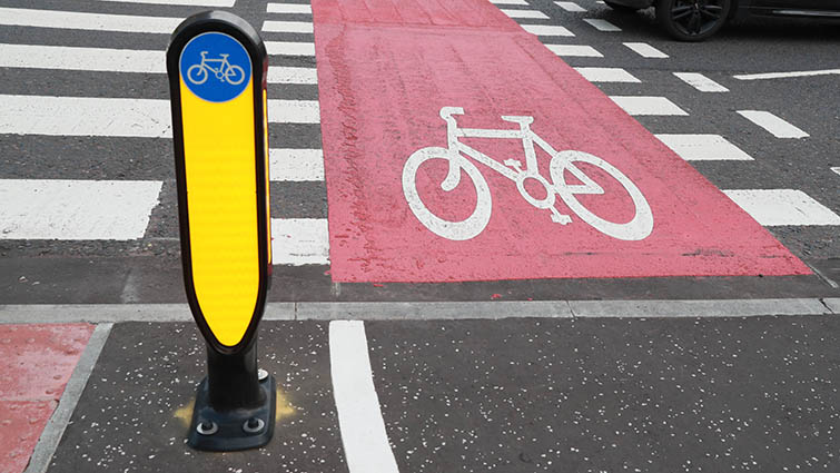 A yellow bollard is in the foreground behind it on the right is a red tarmac cycle lane and on the left a zebra crossing 