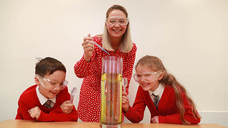 from left to right there is a boy, a Gillian Reilly South Lanarkshire's primary school science development officer and a girl. They are looking at a large beaker containing coloured chemicals. 
all three are wearing red. 