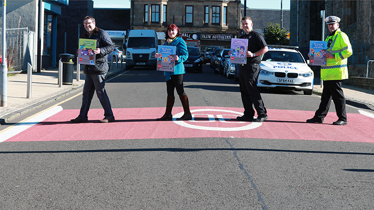 four people walking across a road holding signs for the be smarter than your smartphone campaign. The individual to the right is a uniformed police officer in a Hi Vis coat. 