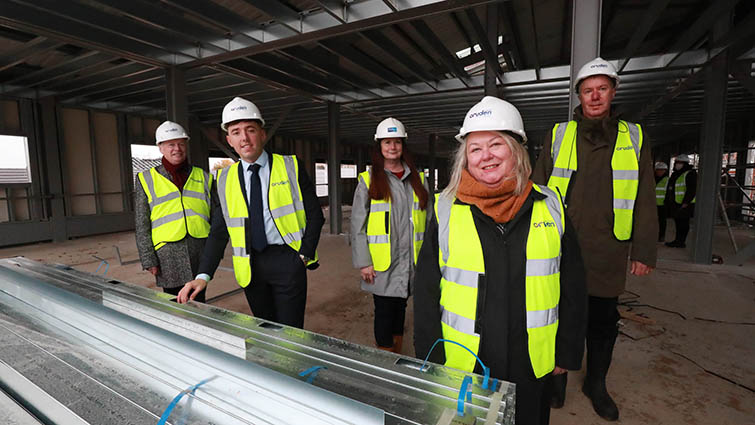 Five people wearing building site PPE stand inside a large building that is still under construction.