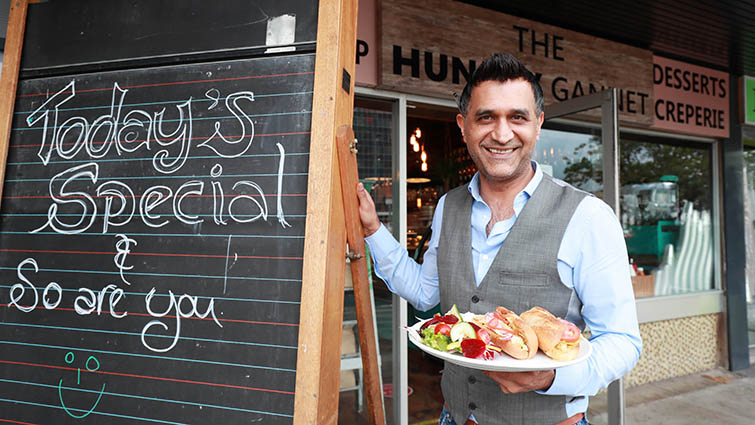 This photo shows Taher Purwaiz the owner of the Hungry Gannet standing in front of the deli, He has a plate of food in his left hand and to his right is a chalkboard on the the message Today's Special and so are you has been written 