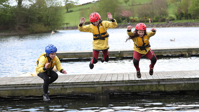 three children wearing wetsuits. lifejackets and helmets are jumping into water from a wooden pontoon 