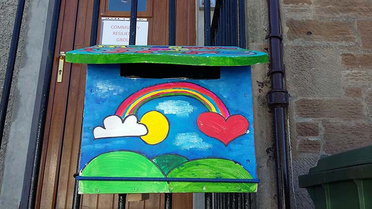 a letterbox attached to iron railings. It has been painted with a cartoon style scene showing a rainbow arching from a yellow sun to a red love heart in a blue sky over green fields