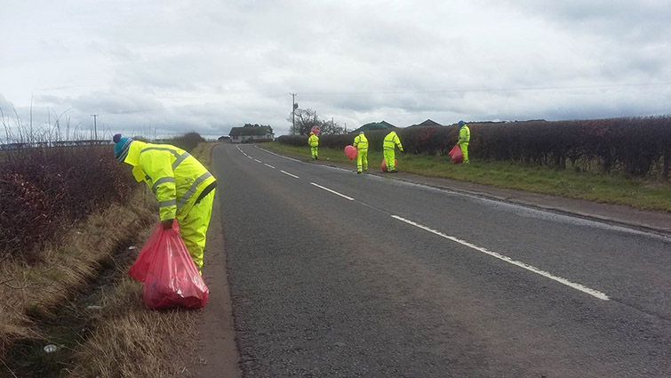 Workers n high visibility jackets are lifting litter on the verges of a roadside in South Lanarkshire. 