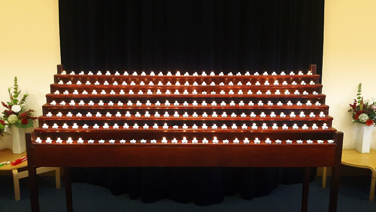 in the centre is the tiered candle holder at South Lanarkshire Crematorium, There is a black curtain behind it and floral displays against white walls to either side. 