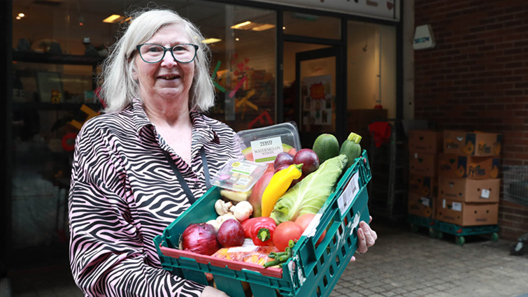 A woman in a grey top is holding a green basket containgin a selection of fresh fruit and vegetables