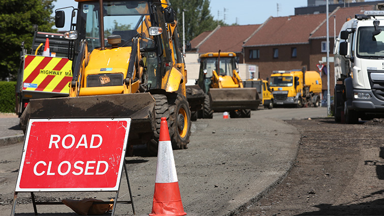 This is a picture showing a number of road maintenance vehicles on a street, in the foreground is a red sign that reads Road Closed and an orange and white traffic cone 