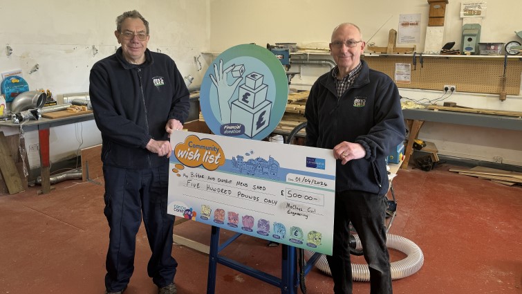 Two members of the Biggar Men's Shed group are standing side by side in their hub holding a big cheque prop