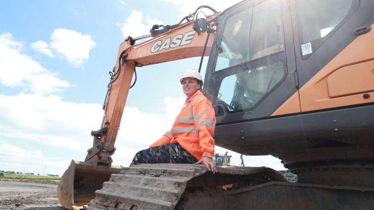 Carly dressed in high visibility jacket and hard hat is sat on the front wheel of a JCB digger looking out across the Blantyre quarry.