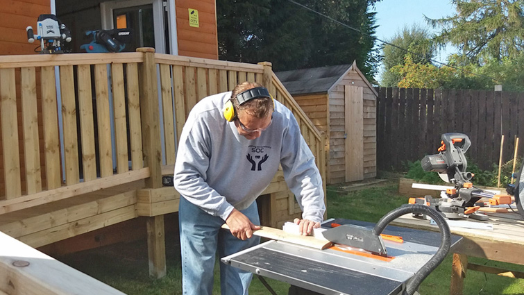 This photo shows a member of the community group working with a sander on a workbench on one of the new planters for the site. He is outdoors in front of the community hub.