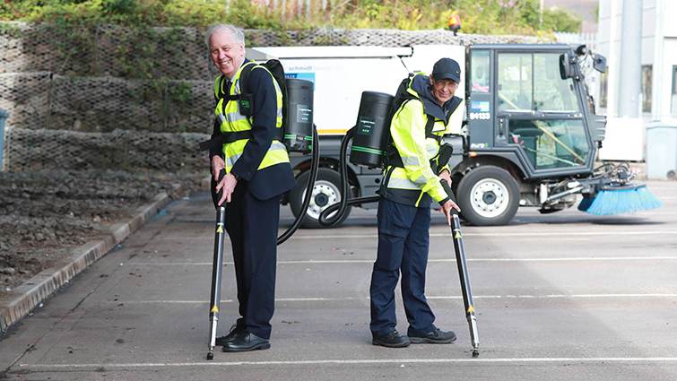 this photo shows Councillor Robert Brown and litter picker Sharon Montgomery standing back to back with the chewing gum machines on their backs, demonstrating them in use.