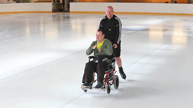 This photo shows care worker Graeme skating with Steven in his wheelchair on EK ice rink. They are looking at the camera.