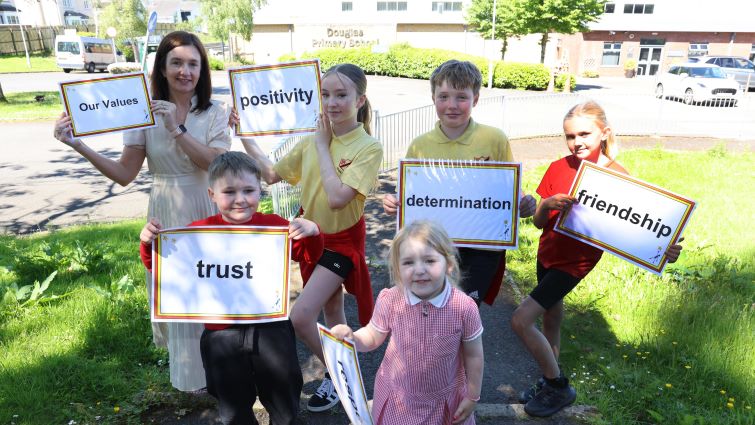 This image shows pupils and a teacher from Douglas Primary School holding up signs with various words including positivity and trust written on them. They are photographed outside the school