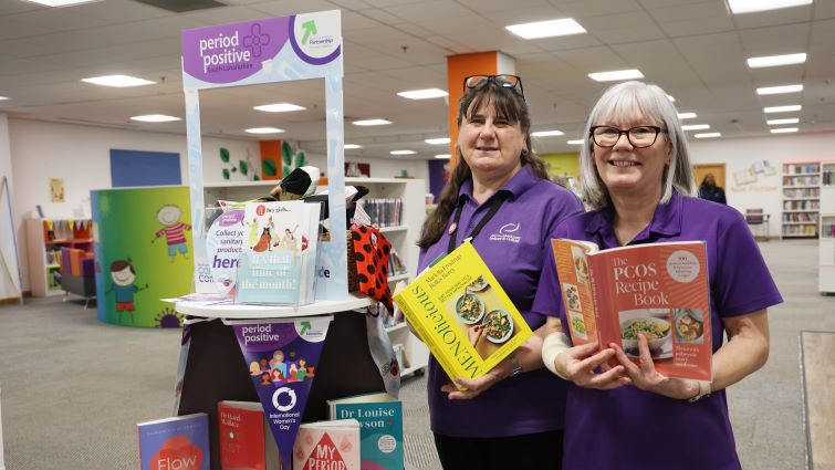 This image shows two librarians holding books which cover a range of topics around women's health, including advice about periods and the menopause, ahead of International Women's Day