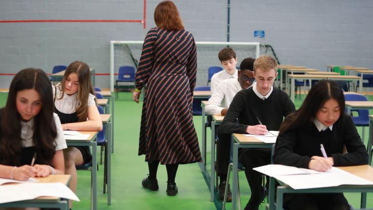 This image shows a number of pupils seated at desks taking a test with the back of a teacher supervising the test
