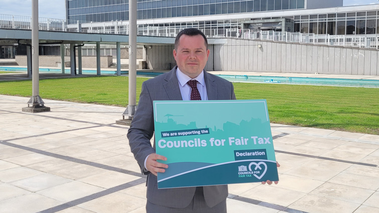 This image shows Councillor Joe Fagan outside council HQ with a sign supporting the council's for fair tax declaration 