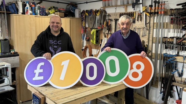 Two male members of the SOC group are pictured in their hub holding large coloured numbers spelling out £1008.
