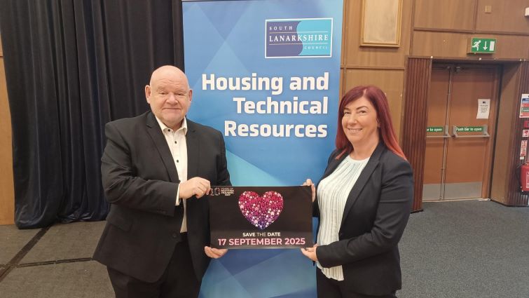 This image shows Councillor Davie McLachlan and Head of Housing Services Sharon Egan holding a poster highlighting Scottish Housing Day 2025 in front of a Housing and Technical Resources pop up banner