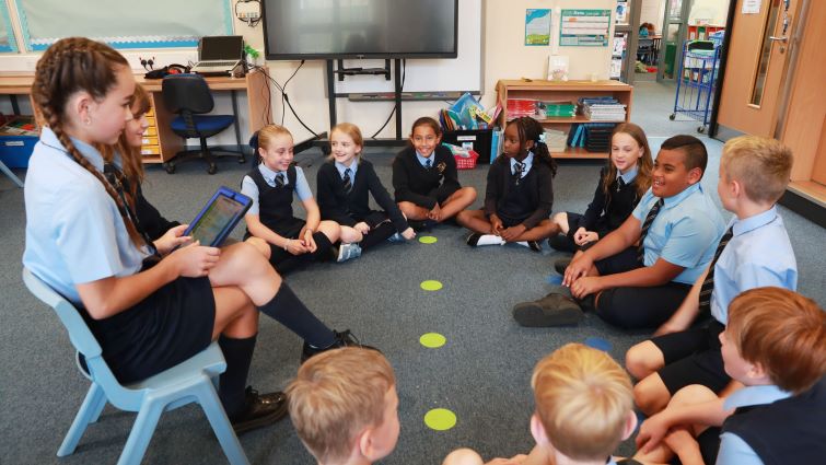 This images shows St Mary's Hamilton pupils sitting in a circle laughing with one child in a chair holding a tablet device