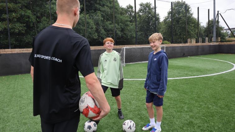 This image shows two young people looking at a sports coach who has his back to the camera and is holding a football. There are two footballs on the ground and the back of the coach's T shirt says head sports coach
