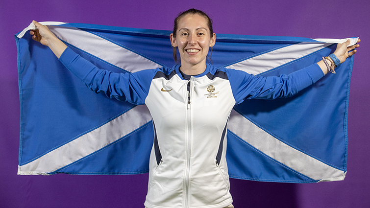 This is an official Team Scotland photo showing Kirsty Gilmour in team colours holding a large saltire behind her. 