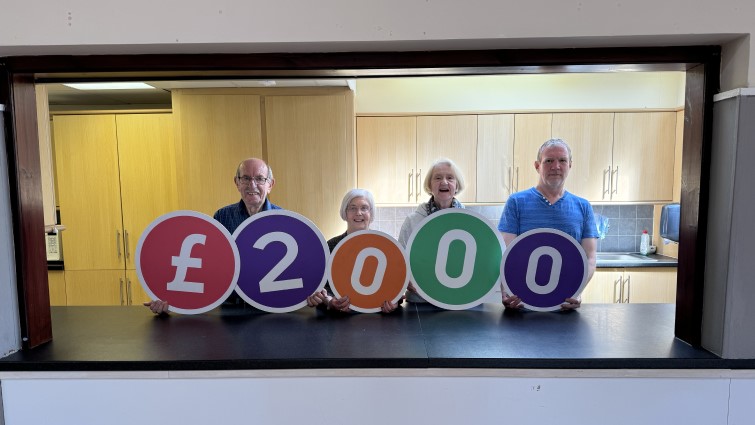 Four of the church volunteer group stand in a line at the kitchen counter holding props which spell out £2000