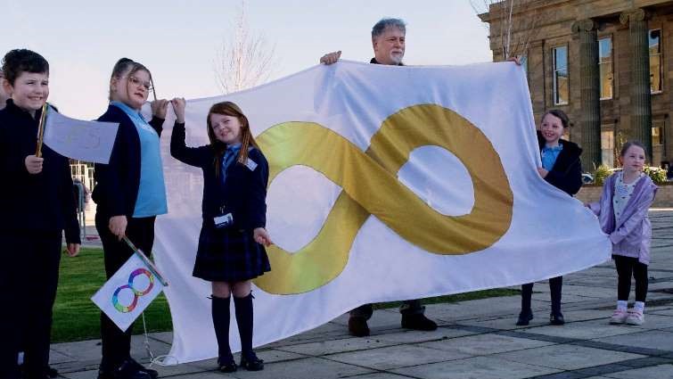 This image shows young people and ARCH Co-ordinator Ramon Hutchingson behind the new Autism Pride gold flag