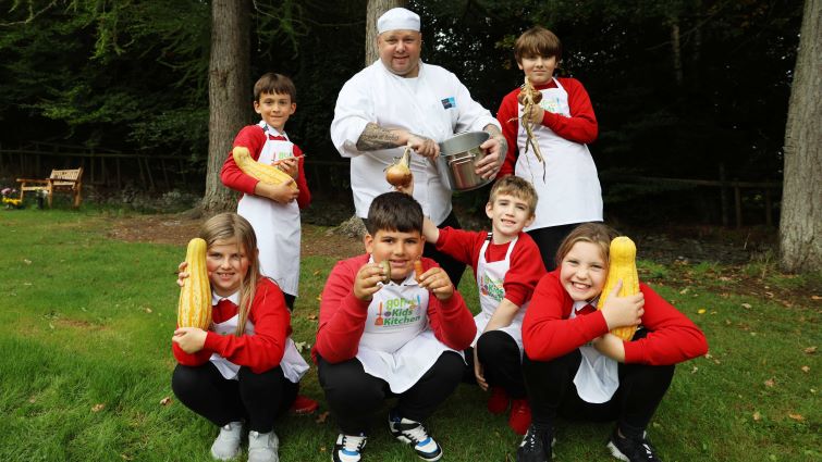 This image shows pupils from Kirkfieldbank primary school holding various vegetable and cooking utensils with Chef Robert, who is stirring a spoon in a pan