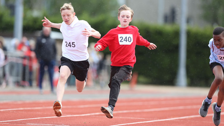This is a picture of two boys crossing the finish line in a 100m race the boy on the left is wearing a white t-shirt and the one on the right is wearing a red long-sleeved top.