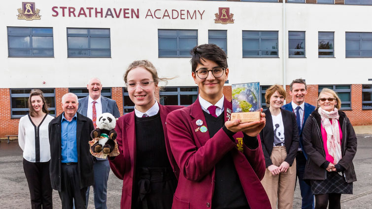 Two Strathaven pupils in uniform stand in the foreground holding the award and the WWF toy panda logo. Lined up behind them are councillor John Anderson, two reps from WWF, Headteacher Kevin Boyd and the council's Julie Richmond. 