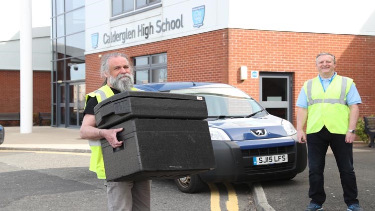 Neil who is bearded and wearing glasses and a hi-vis vest, stands in the foreground holding a box with meals, while Gary, also wearing a hi-vis vest stands behind getting ready to load the council van.