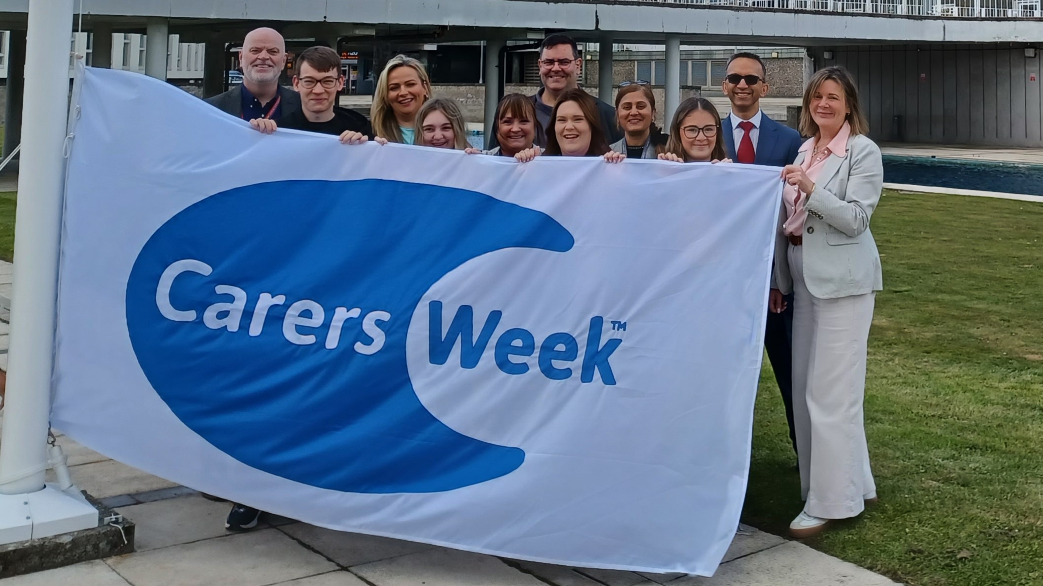 This image shows a range of invited guests holding a flag which says Carers Week