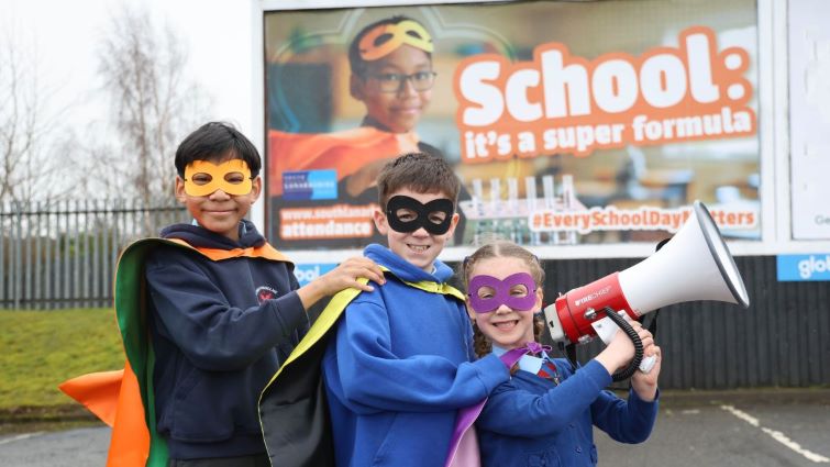 Darius, Ethan and Ellie stand in a line in front of the advert on a billboard, wearing cloaks and eyemasks. Ellie is at the front of the line holding a loudhailer.